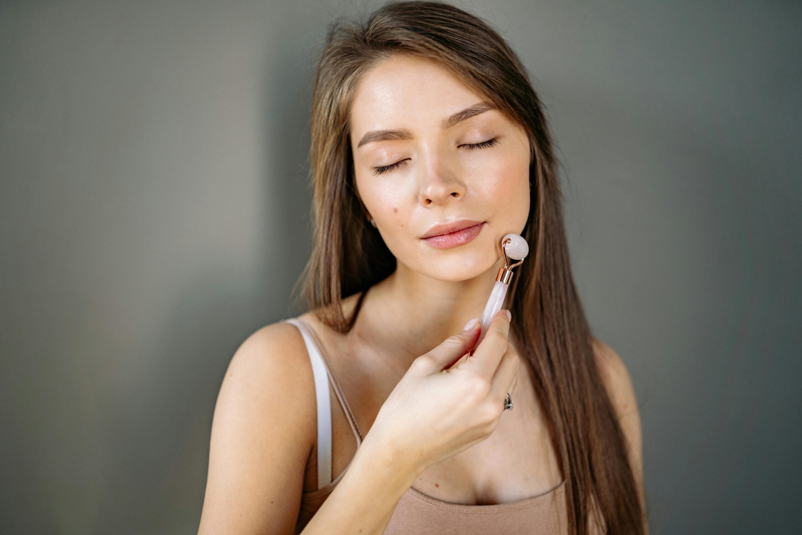 Woman with eyes closed using a jade roller for facial care on gray background.